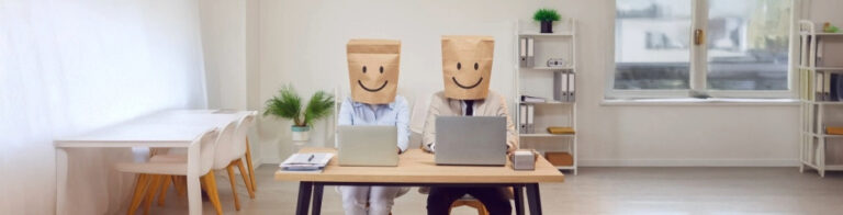 Two people with brown paper bags on their heads sitting at desk in front of laptops.